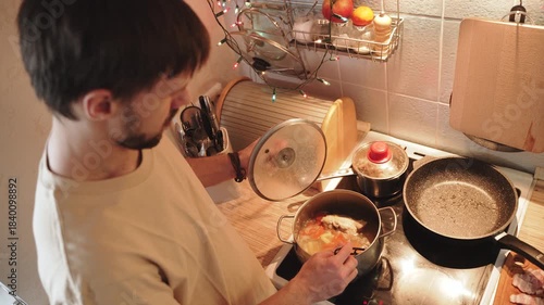 Man opens lid of pot of soup on stove and stirs it with spoon, overhead shot. Male person cooks hot entree with meat in home kitchen, warm light.