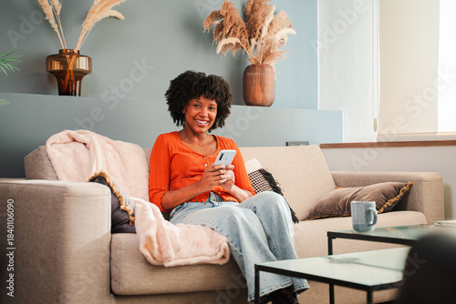 Happy woman using her smartphone while sitting on a soft couch, enjoying her time in a cozy living area filled with decorative plants and a warm atmosphere, engaged in her mobile application.