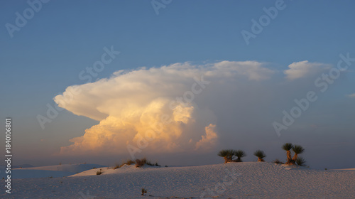 Fototapeta Naklejka Na Ścianę i Meble -  A mesmerizing view of a vast, undulating white sand dune landscape under a serene blue sky, adorned with a massive, golden-hued cumulonimbus cloud at sunset.