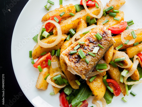 Plate with baked potatoes, piece of fish, vegetables and herbs on the table with tomato sauce.