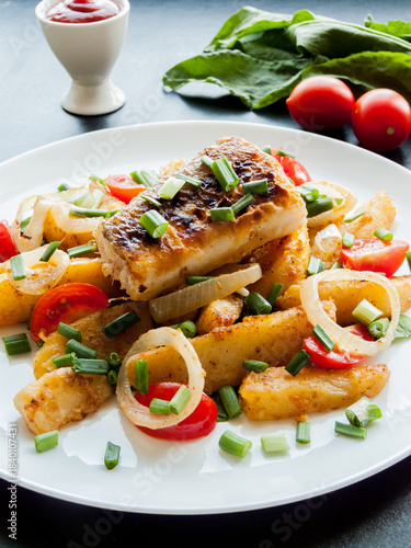 Plate with baked potatoes, piece of fish, vegetables and herbs on the table with tomato sauce.