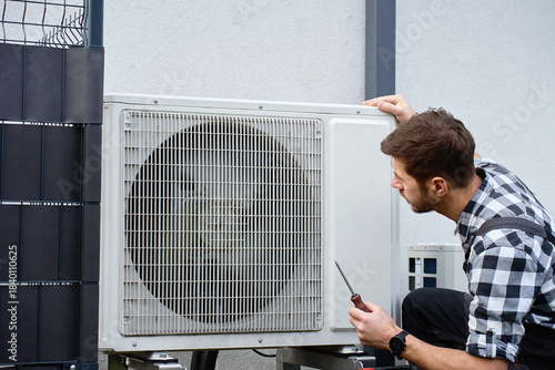 Technician performing maintenance on outdoor heat pump unit near residential building exterior. Man installing air source heat pump, using tools. Concept of HVAC service and heating system repair
