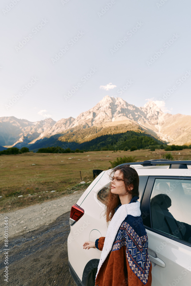 Naklejka premium Woman car mountains road travel landscape: young woman leans on a white SUV beside a mountain road, cozy sweater and soft sunlight over valley and peaks during an outdoor road trip.