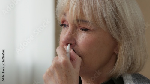 Portrait ill old Caucasian woman spraying nose with nasal spray remedy allergy sneeze senior female retired granny lady unhealthy medicine health care sneezing at home medication allergic symptoms
