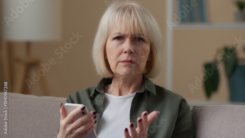 Old retired Caucasian woman with mobile phone sad confused upset looking at camera at home senior female grandmother lady with smartphone bad news reject notification feeling negative connection error