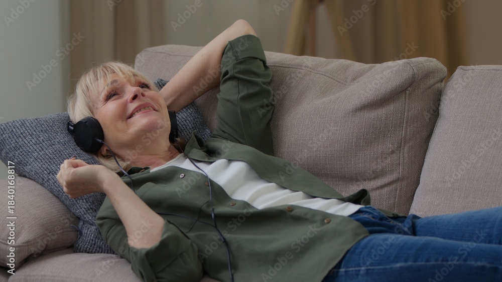 Obraz premium Relaxed old Caucasian woman listening music in headphones enjoying song moving hands dancing lying on couch sofa happy carefree grandmother senior female retired lady listen musical track at home