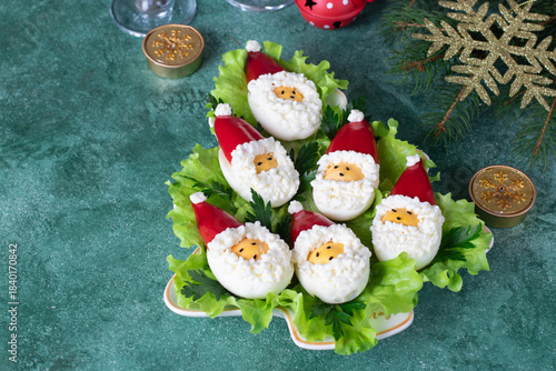Festive deviled eggs in the shape of Santa Claus on lettuce leaves on Christmas tree-shaped plate on green background