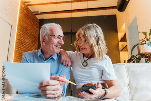 Elderly couple happily managing household finances and planning future retirement, reviewing important documents and bills together in their comfortable home