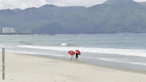 Two people with red umbrellas walk on a picturesque beach in Cam Ranh, Vietnam, with mountains and gentle waves. Concept of coastal vacation.