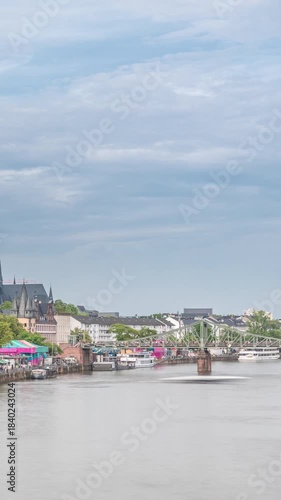 Aerial hyperlapse of pleasure boats on the Rhine Main, framed by the spire of Kaiserdom St. Bartholomaus. Eiserner Steg bridge and Ferris wheel add vibrancy to Frankfurt's scenic waterfront timelapse