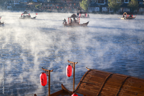 Serene, tranquil morning in traditional Chinese water town. Misty fog rises from river as people enjoy peaceful ride on an ancient wooden boat with red lanterns