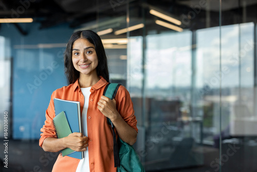 Portrait of a young smiling Indian female student standing indoors, holding a backpack and books, looking at the camera