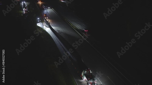 Spectacular heavy transport of a wind turbine rotor blade at night on a multi-lane highway. The oversized truck is illuminated by escort vehicles on the dark road.