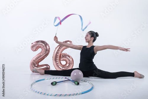 Rhythmic gymnast girl performs a ribbon twirling move, with other rhythmic gymnastics equipment and a number 26 balloon placed next to her. Celebrate New Year 2026, white background