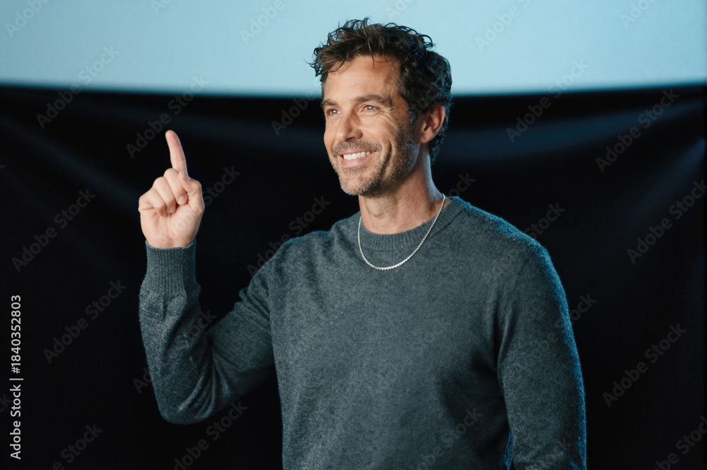 Fototapeta premium Man in gray sweater pointing upward with index finger while smiling against dark background