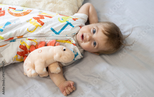 funny little girl with a dinosaur toy in a sleeping bag woke up lying on her bed at home. Relaxing comfortably in the bedroom. The concept of purity, innocence, and tender moments of childhood