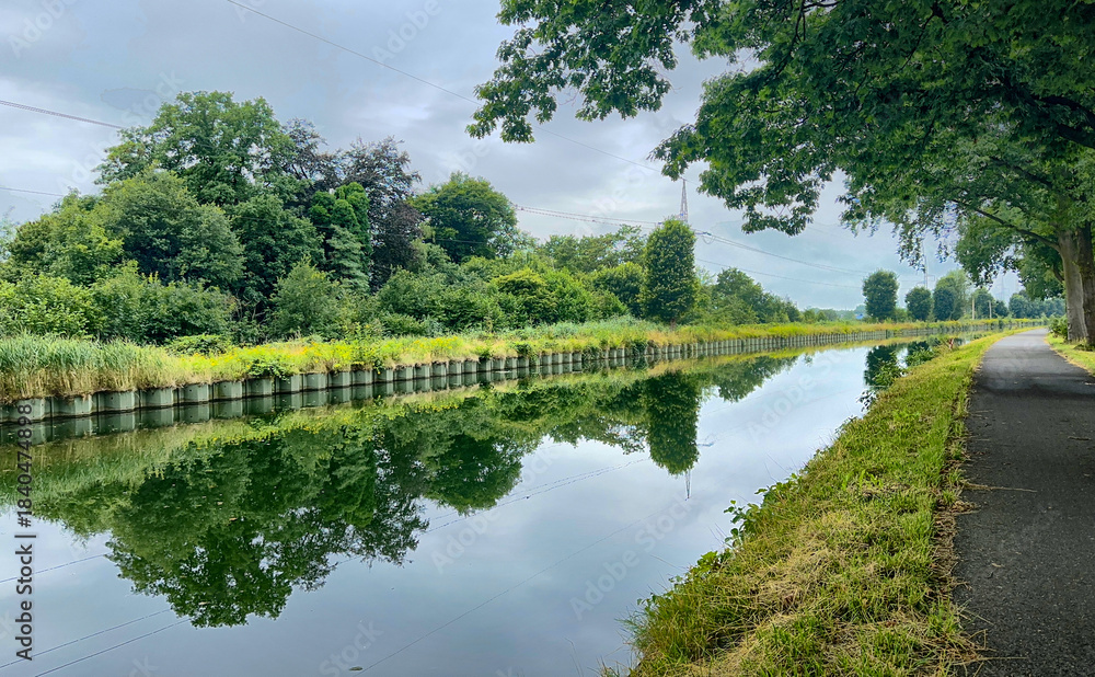 Fototapeta premium Calm canal reflecting tree-lined path with smooth mirror water and paved towpath, quiet morning scene ideal for cycling