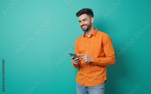 Full length photo of pretty cute guy wear orange shirt typing modern device pointing empty space isolated teal color background. High quality