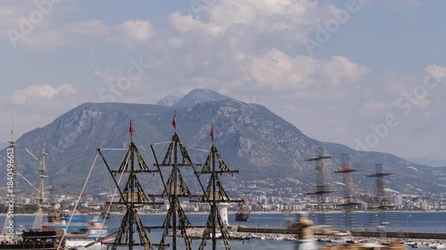 A historical ship replica stands prominently in the foreground of a bustling harbor scene at Alanya, with the city and mountains visible in the background.