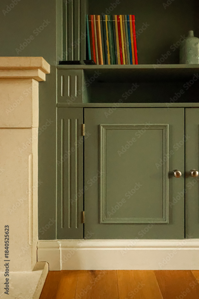 Obraz premium Green painted custom built in cabinet and bookshelf with small colorful books next to a decorative stone fireplace