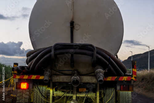 Mauritius - Rear view of a water tanker truck on a rural dirt road at dusk, with coiled hoses, and a natural landscape in the background