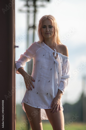 Young woman with long blonde hair wearing a white blouse poses confidently against a rust-colored structure, showcasing a relaxed summer vibe and natural beauty