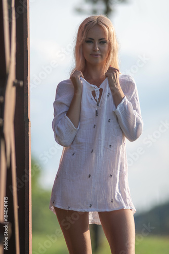 Young woman with long blonde hair wearing a white blouse poses confidently against a rust-colored structure, showcasing a relaxed summer vibe and natural beauty