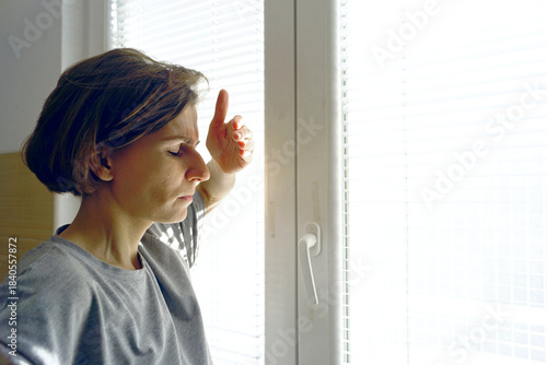Woman shielding eyes from window light during migraine attack with photophobia. Symbolizes light sensitivity, visual trigger avoidance, photosensitive response, and optical discomfort protection.