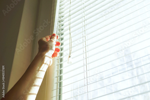 Hand adjusting white venetian blinds to reduce light exposure for migraine photophobia relief. Illustrates light control strategy, brightness reduction, sensory trigger elimination, and visual comfort