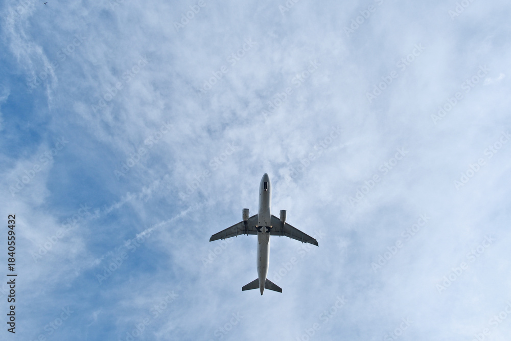 Fototapeta premium White Plane Overhead in Light Clouds. White Jet Soaring Overhead in Clear Blue Sky with Light Clouds