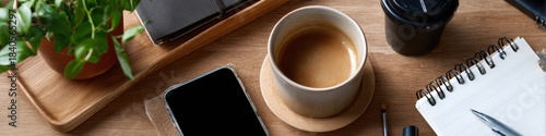 Office desk with coffee, notepad, pen, smartphone, potted plant, and planner on wooden surface