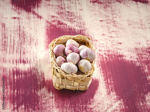 A tiny woven basket overflows with bulbs of vibrant garlic. The scene is set upon a mottled purple painted wood surface, bathed in gentle illumination