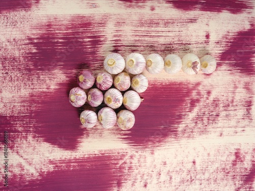 A formation of garlic bulbs creates a directional arrow on a textured and painted wood surface. The purple background provides a striking contrast