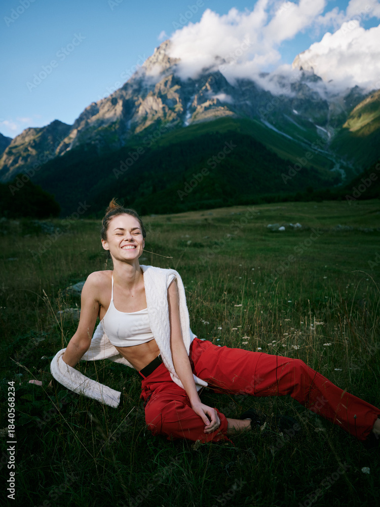 Naklejka premium Adventure in a meadow with towering mountains, a smiling woman sits on grass after a workout, bright red pants, white top, towel over shoulder, natural scenery