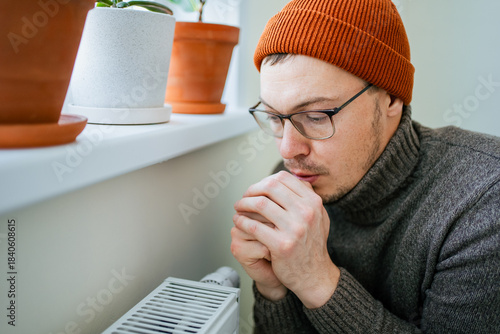 Man wearing warm hat and sweater suffering from cold at home, paying expensive heating bills and saving money during energy crisis, warming hands near radiator due to high costs