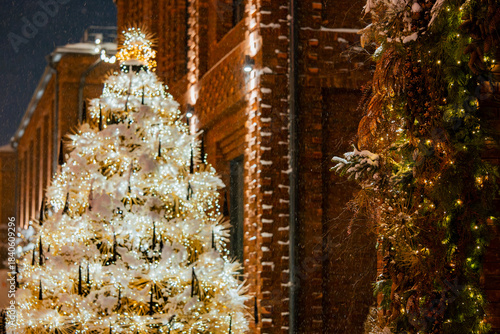 Brightly lit christmas tree sparkling with warm white lights and draped in snow stands outside a brick building, creating a festive winter scene during a snowing holiday evening