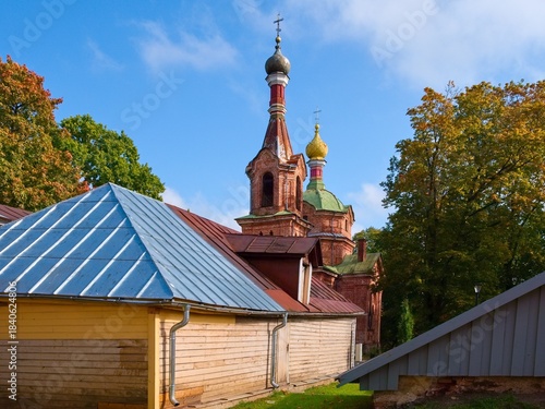 Tableau sur toile Old brick Orthodox church with colorful onion domes rising above wooden village houses and autumn trees under blue sky, traditional Russian architecture and countryside landscape