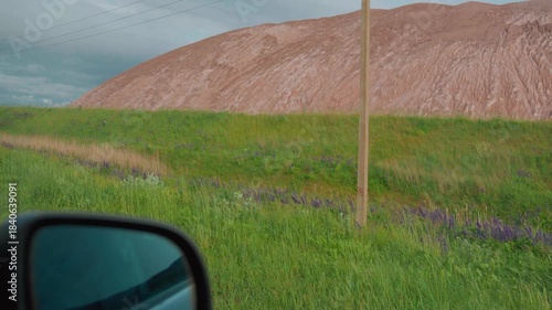 Personal perspective driving along a quiet rural road, passing a large industrial slag heap from a mining operation under a bright sky, illustrating travel through a modified landscape