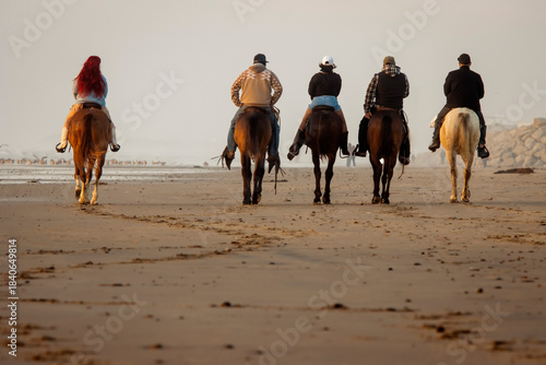 Behind shot of five people, men and women, riding horses on a beach