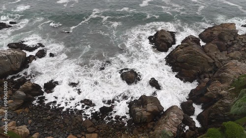 Dramatic aerial reveal from coastal vegetation to stormy ocean waves hitting rocks in fog.
