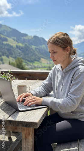 Concentrated young Swiss woman typing on her laptop on a sunny mountain terrace with a scenic alpine view in the background. Modern remote work and flexible workplace concept in nature.
