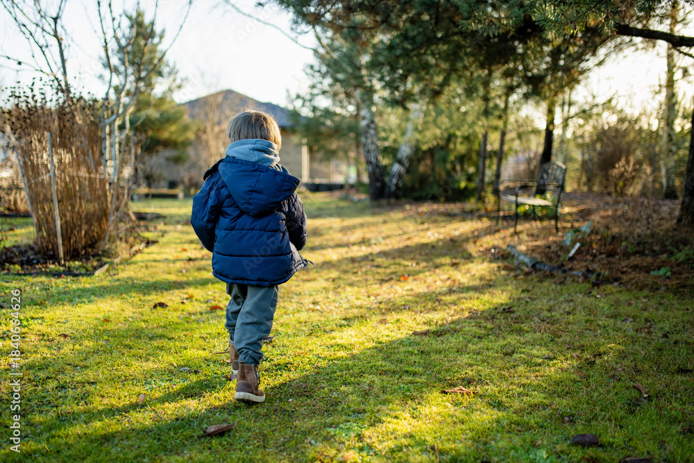Obraz premium Smiling child in hoodie and puffer jacket stands in a green garden on a grey day. Mild European winter or late autumn, quiet moment between seasons.