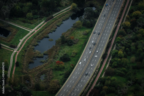 Aerial view of the sleek multi-lane highway cuts through a tapestry of green and gold foliage alongside a dark, tranquil canal, Warsaw, Poland.