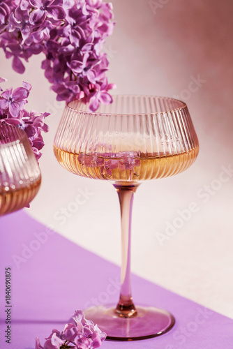 Elegant floral cocktail with lilac blossoms inside vintage coupe glass, placed on a pastel purple background with bouquet details