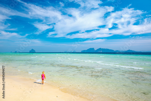 Child standing at the edge of a calm. Clear ocean on a tropical beach. Observing the water and distant seascape with small islands under a blue sky. Enjoying a peaceful moment during a summer vacation