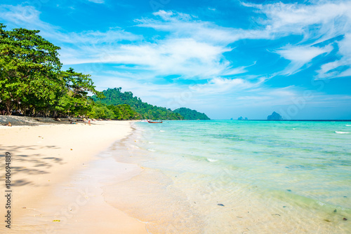 White sand tropical beach meeting clear turquoise water and lush green jungle, featuring a traditional longtail boat anchored offshore with distant islands under a bright blue sky