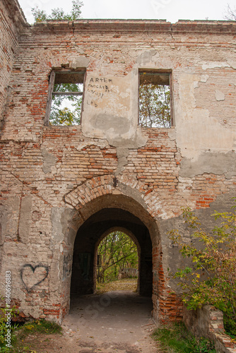 Weathered brick archway passage through deteriorating building facade with broken windows, peeling plaster, graffiti and overgrown vegetation in autumn