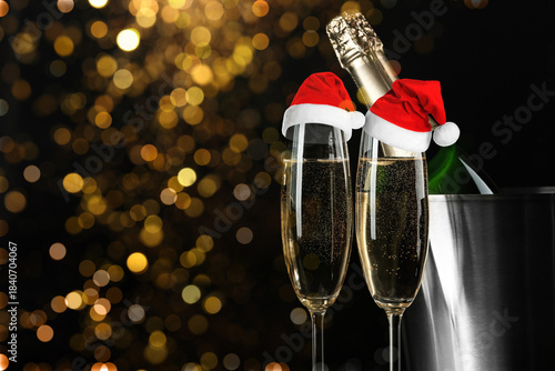 Sparkling wine in glasses with small Santa hats against blurred lights on black background