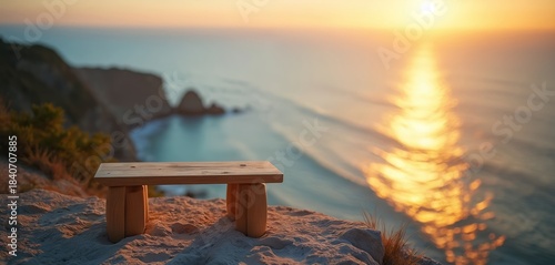 Wooden stool sits on cliff edge overlooking calm sea at sunset. Ocean reflects golden sun rays. Peaceful coastal landscape background. Serene natural view.