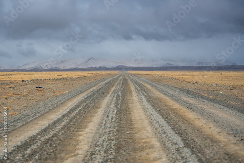 Gravel road stretching from Altai towards the Mongolian border with snowy mountain ridge. Desert steppe landscape under stormy cloudy sky showcasing wild remote terrain